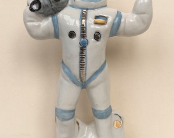 May include: A white porcelain figurine of an astronaut wearing a blue and white spacesuit. The astronaut is holding binoculars and has a surprised expression on their face. The figurine is standing on a white base with gold accents.