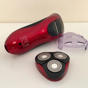 May include: A red electric razor with a black and silver rotary head and a clear plastic protective cover. The razor has the text "HK-4803 220V-240V 10 min" printed on the side.