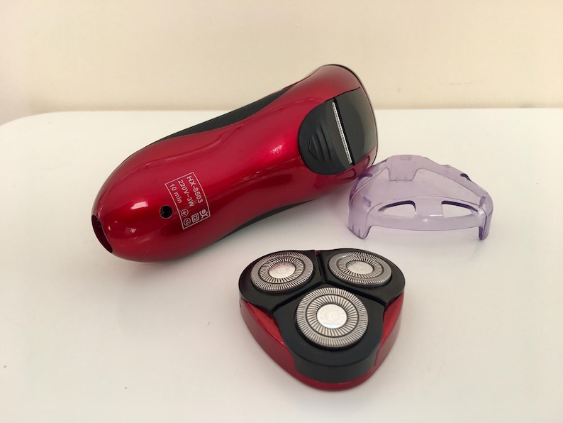 May include: A red electric razor with a black and silver rotary head and a clear plastic protective cover. The razor has the text "HK-4803 220V-240V 10 min" printed on the side.