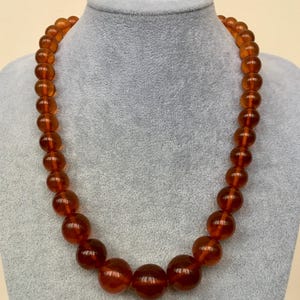 Puede incluir: Un collar largo hecho de grandes cuentas redondas de color &aacute;mbar. Las cuentas son todas del mismo tama&ntilde;o y est&aacute;n enhebradas en un cord&oacute;n delgado y transparente. El collar se muestra en un maniqu&iacute; gris.