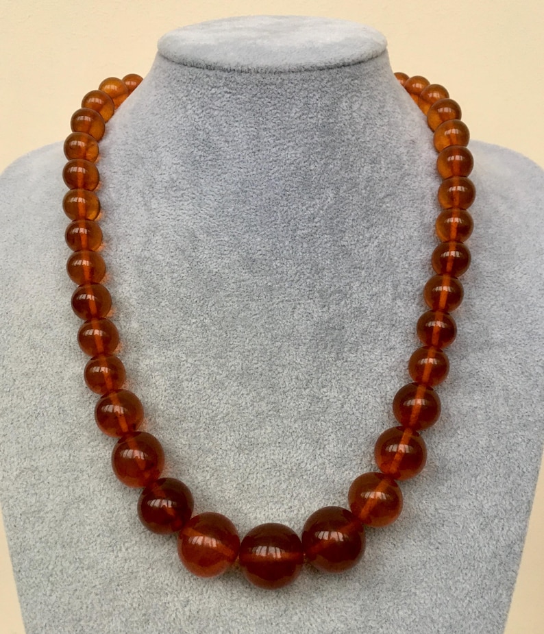 Puede incluir: Un collar largo hecho de grandes cuentas redondas de color &aacute;mbar. Las cuentas son todas del mismo tama&ntilde;o y est&aacute;n enhebradas en un cord&oacute;n delgado y transparente. El collar se muestra en un maniqu&iacute; gris.