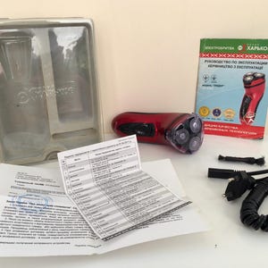 May include: A red electric shaver with a rotating head and a black cord. The shaver is packaged in a clear plastic box with instructions and a warranty card. The instructions are in Ukrainian and Russian.