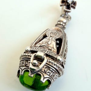 May include: A silver pendant featuring an intricate cage design with a central figure in relief, Cyrillic text engravings, and a rounded, translucent green stone at the base. The top includes a small cross-shaped bail with a clear inset stone.