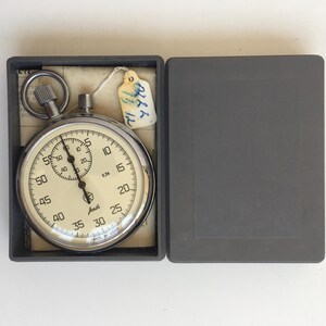 May include: A vintage stopwatch with a cream-colored face, black numerals, and a silver metal casing. The stopwatch is in a gray case with the lid open. A small tag is attached to the stopwatch.