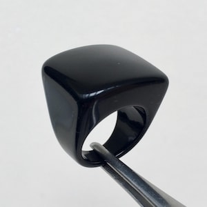 May include: A sleek, black ring with a square face and curved sides. The ring is held by silver tweezers, highlighting its polished surface. The design is contemporary and minimalist. The ring's measurements are in centimeters.