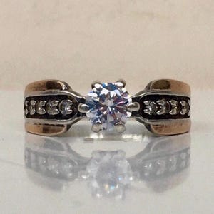 May include: A silver ring with a large, clear, round gemstone at the center. The band features gold-colored accents and small, clear stones. The ring is set against a reflective surface.