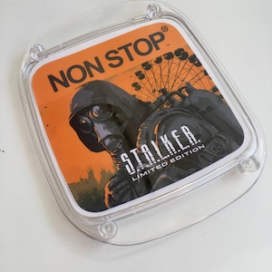 Peut inclure: Un étui en plastique transparent avec un insert imprimé. L'insert affiche le texte "NON STOP" et "STALKER LIMITED EDITION". Le design présente une figure avec un masque à gaz sur fond orange, avec une grande roue.