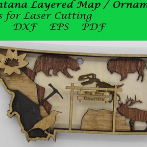 Könnte beinhalten: Ein mehrschichtiges Holz-Ornament einer Karte von Montana mit einem Braunbären, einem Bison, einem Dinosaurierskelett, einer Spitzhacke, einem Vogel, einer Blume und einem Elch. Das Ornament hat ein Schild mit der Aufschrift "Big Sky Country".