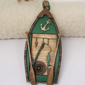 May include: Wooden ornament shaped like a green canoe with brown oars, a fishing rod, a fish, and an anchor. The ornament has a wood grain pattern and is decorated with a green and brown color scheme.