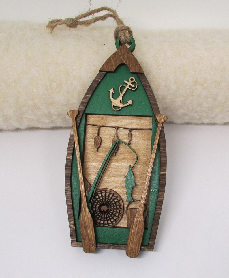 May include: Wooden ornament shaped like a green canoe with brown oars, a fishing rod, a fish, and an anchor. The ornament has a wood grain pattern and is decorated with a green and brown color scheme.