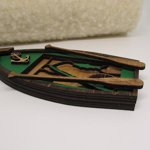 May include: Wooden ornament shaped like a boat with green interior and brown exterior. The boat has two oars and an anchor inside. The ornament has a string attached to the top for hanging.