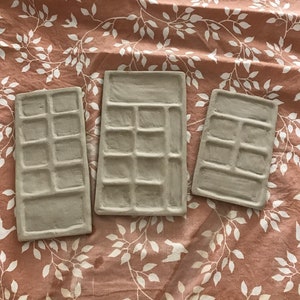 Puede incluir: Tres bandejas rectangulares de cerámica con múltiples compartimentos cuadrados. Las bandejas son de color marrón claro y están dispuestas sobre una tela floral rosa y blanca.