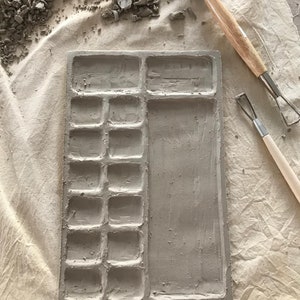 Puede incluir: Una paleta de cerámica gris con 12 pozos individuales y un pozo rectangular más grande. La paleta está sobre una superficie de tela blanca con algunos restos de arcilla esparcidos alrededor. También se pueden ver dos herramientas de escultura.
