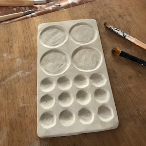 Puede incluir: Una paleta de pintura de cerámica blanca con 19 pozos circulares para mezclar pintura. La paleta está sobre una superficie de madera.