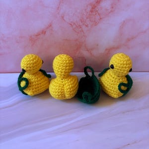 May include: Four handmade crochet items: three yellow ducklings with green accents and a green bag. The ducklings have black eyes. The items are arranged on a pink and white marble-patterned surface.