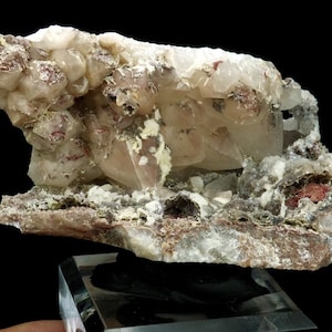 May include: A natural mineral specimen featuring a cluster of translucent, light-coloured crystals with reddish and brown inclusions. The mineral sits on a dark, textured base, and is displayed on a clear stand against a black background.