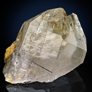 May include: A large, clear quartz crystal with multiple facets and internal striations. The crystal has a light, translucent appearance with some golden and black inclusions. The overall shape is angular and geometric, showcasing natural mineral formations.