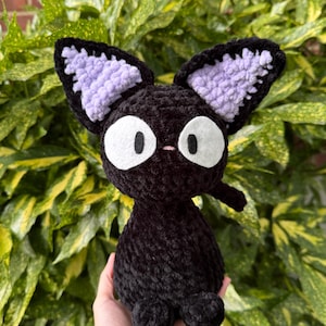 Puede incluir: Un peluche de gato negro hecho a mano con ganchillo, con ojos blancos grandes, una pequeña nariz rosa y orejas internas de color lavanda. El gato tiene una pequeña cola y se sostiene en una mano. El peluche está sobre un fondo de follaje verde y amarillo.