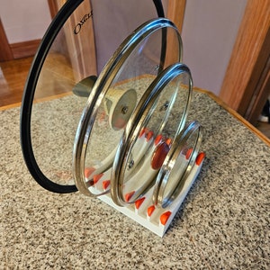 May include: A white plastic lid organizer with four slots holds four glass lids. The lids are stacked on top of each other, with the largest lid on the bottom and the smallest lid on top. The largest lid has a black rim and the text "OXO" printed on it.