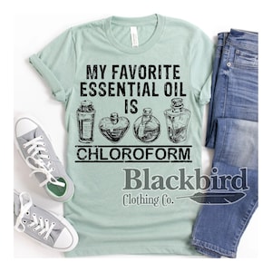 May include: A light green t-shirt with the text "MY FAVORITE ESSENTIAL OIL IS CHLOROFORM" and illustrations of four apothecary bottles. The shirt is paired with gray sneakers and blue jeans. The Blackbird Clothing Co. logo is at the bottom.