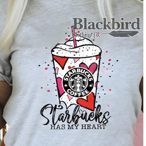 May include: A light gray t-shirt featuring a Starbucks coffee cup design. The cup is decorated with red and pink hearts, and the Starbucks logo is visible. The text "Starbucks HAS MY HEART" is written below the cup. The brand name "Blackbird Clothing Co." is at the top.