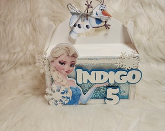 May include: A white and blue favor box with a Frozen theme. The box features a picture of Elsa, a snowflake, and the text "INDIGO 5".