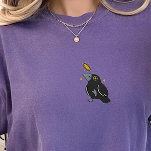 Puede incluir: Camiseta morada con un diseño bordado de un cuervo negro con una moneda de oro. La palabra "GRAPE" está en un círculo morado. Parte de la colección Pinespice Embroidered & Cozy. Una prenda casual y cómoda.