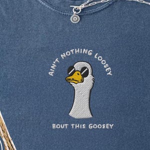 Puede incluir: Una camiseta azul vaquera con la frase bordada "Ain't Nothing Loosey Bout This Goosey" y un dibujo de un ganso con gafas de sol. Un collar plateado con un colgante descansa sobre la camiseta. La camiseta está doblada sobre una superficie de mezclilla clara.