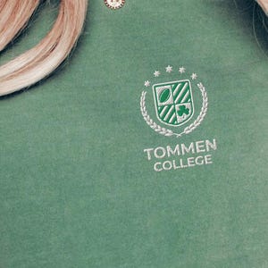 May include: Light green t-shirt with the text "TOMMEN COLLEGE" and a crest design. The crest features a shamrock, a shield, and stars. The shirt is a solid color and has short sleeves. The words "LIGHT GREEN" are in a circle at the bottom.