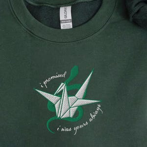 May include: Forest green sweatshirt with embroidered origami crane design. The crane is white and gray, surrounded by a green snake-like design, with the text "I promised I was yours always". The sweatshirt is cozy and embroidered.