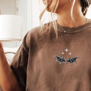 May include: A brown t-shirt featuring a black bat and stars graphic. The t-shirt is made from a soft, comfortable material and has a relaxed fit.