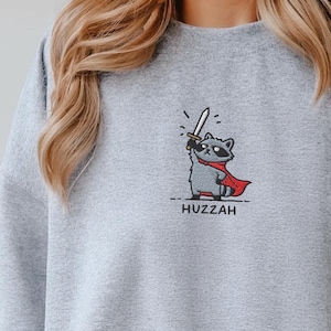 Könnte beinhalten: Sportgraues Sweatshirt mit einem gestickten Cartoon-Waschbären, der ein Schwert hält und einen roten Umhang trägt. Das Wort "Huzzah" ist darunter gedruckt. Das Sweatshirt hat den Markennamen "Pinespice" auf der oberen linken Seite.
