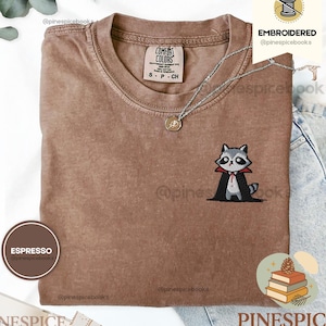May include: An espresso-colored t-shirt featuring a small embroidered graphic of a raccoon dressed as a vampire in a black cape with a red collar. The shirt has a crew neckline and a Comfort Colors brand label on the inside of the collar.