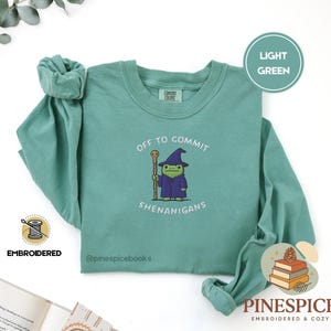 Puede incluir: Camiseta verde claro de manga larga con el texto "OFF TO COMMIT SHENANIGANS" y un personaje de dibujos animados de mago. La camiseta está bordada y presenta la marca "Pinespice".