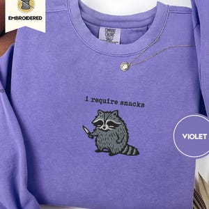May include: Violet crewneck sweatshirt with embroidered raccoon graphic holding a knife. The text "i require snacks" is above the raccoon. The sweatshirt is made of 100% cotton and is pre-shrunk.
