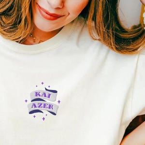 Może przedstawiać: Biała, oversize'owa koszulka z haftowanym wzorem w kolorze fioletowym, na którym widnieje napis "KAI AZER". Koszulkę nosi osoba siedząca na dywanie i czytająca książkę.