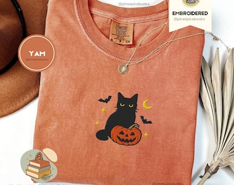 May include: An orange Comfort Colors t-shirt with an embroidered Halloween design. The design features a black cat sitting on a carved pumpkin, with bats and a crescent moon. A gold necklace is draped across the shirt.