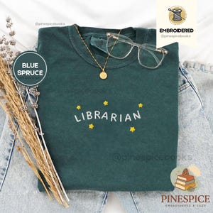 Puede incluir: Camiseta azul abeto con la palabra "LIBRARIAN" bordada en blanco, rodeada de estrellas amarillas. La camiseta se combina con gafas de montura transparente y un collar dorado. El texto "BLUE SPRUCE" está en un círculo a la izquierda.