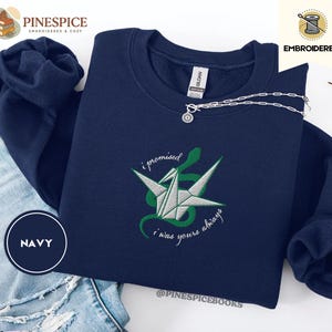K&ouml;nnte beinhalten: Marineblaues Sweatshirt mit gesticktem Origami-Kranich- und Schlangen-Design. Der Text "i promised i was yours always" ist in einer Schreibschrift gestickt. Eine silberne Halskette mit Anh&auml;nger liegt auf dem Sweatshirt.