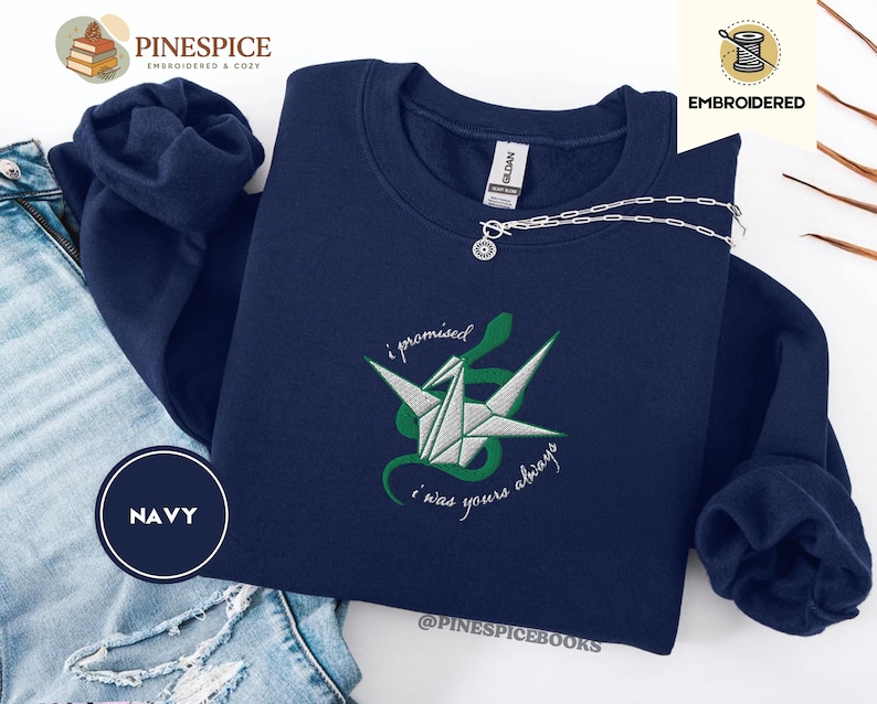 K&ouml;nnte beinhalten: Marineblaues Sweatshirt mit gesticktem Origami-Kranich- und Schlangen-Design. Der Text "i promised i was yours always" ist in einer Schreibschrift gestickt. Eine silberne Halskette mit Anh&auml;nger liegt auf dem Sweatshirt.