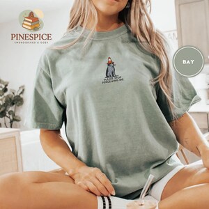 May include: Sage green t-shirt with a graphic of a cat wearing a traffic cone and the text "PLEASE STOP PERCEIVING ME." The shirt is a relaxed fit, short-sleeved style.