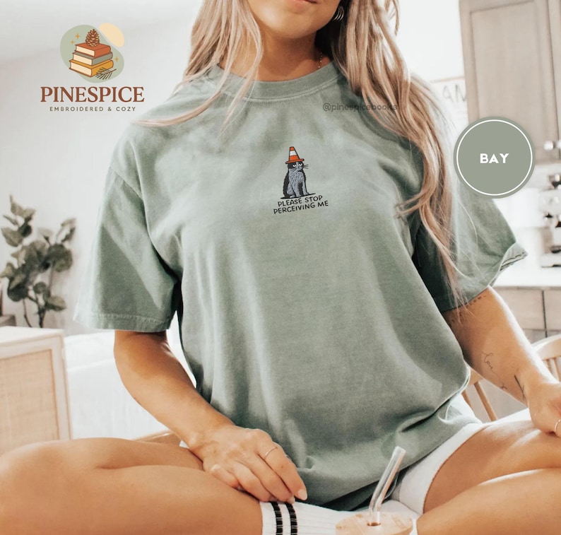 May include: Sage green t-shirt with a graphic of a cat wearing a traffic cone and the text "PLEASE STOP PERCEIVING ME." The shirt is a relaxed fit, short-sleeved style.