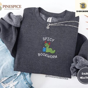 Puede incluir: Sudadera gris jaspeado oscuro con el texto bordado "Spicy Bookworm" y un personaje de gusano verde con gafas y un libro. La sudadera tiene cuello redondo y mangas largas. Una cadena de plata est&aacute; drapeada en la parte superior.