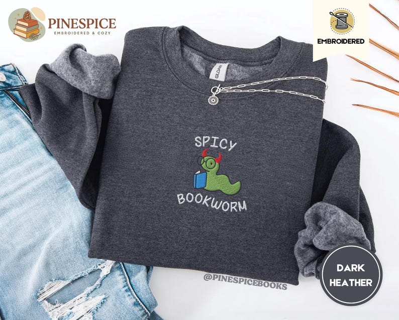 Puede incluir: Sudadera gris jaspeado oscuro con el texto bordado "Spicy Bookworm" y un personaje de gusano verde con gafas y un libro. La sudadera tiene cuello redondo y mangas largas. Una cadena de plata est&aacute; drapeada en la parte superior.