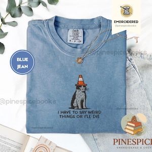 May include: Blue jean-colored t-shirt with an embroidered cat wearing a traffic cone hat. The shirt has the text "I HAVE TO SAY WEIRD THINGS OR I'LL DIE." A Comfort Colors tag is visible. A gold necklace is draped on the shirt.