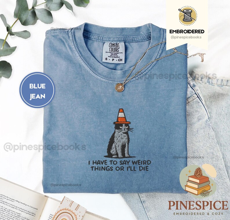May include: Blue jean-colored t-shirt with an embroidered cat wearing a traffic cone hat. The shirt has the text "I HAVE TO SAY WEIRD THINGS OR I'LL DIE." A Comfort Colors tag is visible. A gold necklace is draped on the shirt.