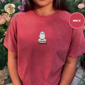May include: Brick-red t-shirt with a small embroidered owl design sitting on books. The owl has large, round eyes. The shirt has short sleeves and a crew neck. The word 'BRICK' is in a red circle.