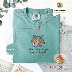 May include: Seafoam green t-shirt with an embroidered cat wearing yellow sunglasses. The cat is gray with purple whiskers. The text "what have I done with my lives?" is below the cat. The shirt is from Comfort Colors.