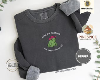 May include: Pepper-colored sweatshirt with embroidered frog wearing a pink cowboy hat. Text reads "Hang on partner, I'm overstimulated." Made of 100% cotton. Features the brand name "Pinespice" and the word "Embroidered".