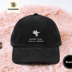 May include: Black corduroy baseball cap with the text "DEFEND YOUR PUBLIC LIBRARY" embroidered in white. The cap features a design of a person on a horse. The word "BLACK" is in a black circle.